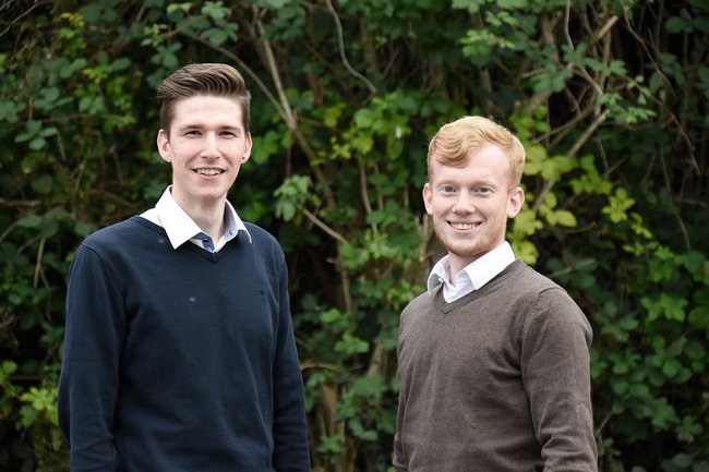 Die 2 jungen Teammitglieder stehen vor einer Hecke und lächeln in die Kamera