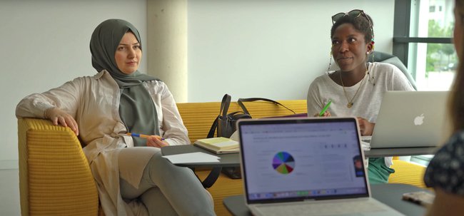 Auf einer Couch sitzt links eine Frau mit Kopftuch, rechts eine schwarze Frau. Im Vordergrund sitzt eine Person, die man nur angeschnitten sieht vor einem aufgeklappten Laptop - die Personen sprechen miteinander
