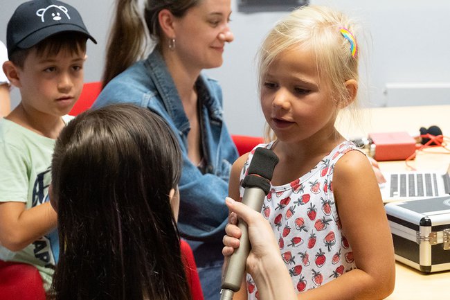 Eine junge Frau hält einem Mädchen ein Mikrofon hin, damit sie etwas sagen kann - im Hintergrund eine junge Frau am Laptop und ein Junge mit Mütze