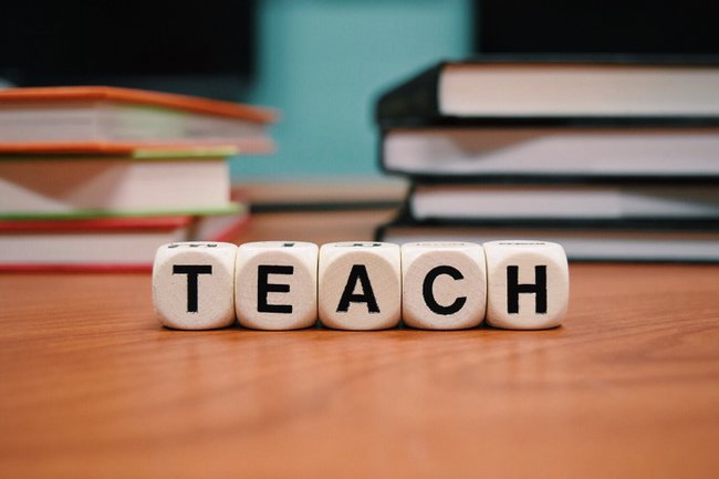 Symbolbild: auf dem Schreibtisch sind mit Würfeln die Worte "Teach" geformt, im Hintergrund sind Bücher sichtbar