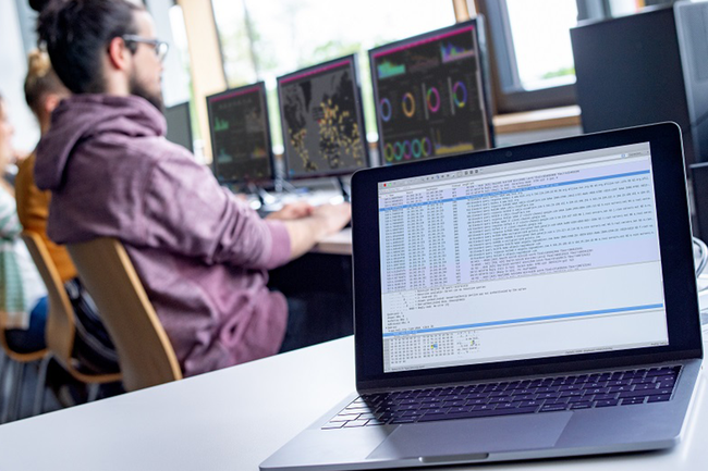 Symbolbild: Im Vordergrund ist ein Laptop mit Daten sichtbar, weiter hinten sitzen Studierende an Bildschirmen, auf denen ebenfalls Daten sichtbar sind