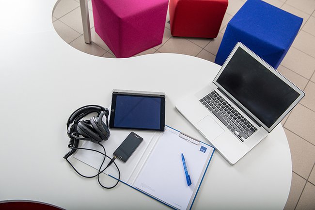 Auf einem Tisch liegen ein Laptop, ein Handy, ein Tablet, ein Kopfhörer und ein Notizbuch - hinter dem Tisch stehen Sitzwürfel