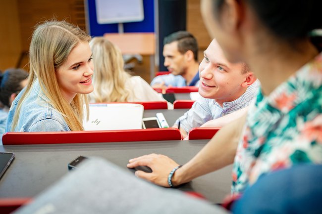 Im Hörsaal sind mehrere Studierende sichtbar, die fröhlich auf ihre nächste Vorlesung warten