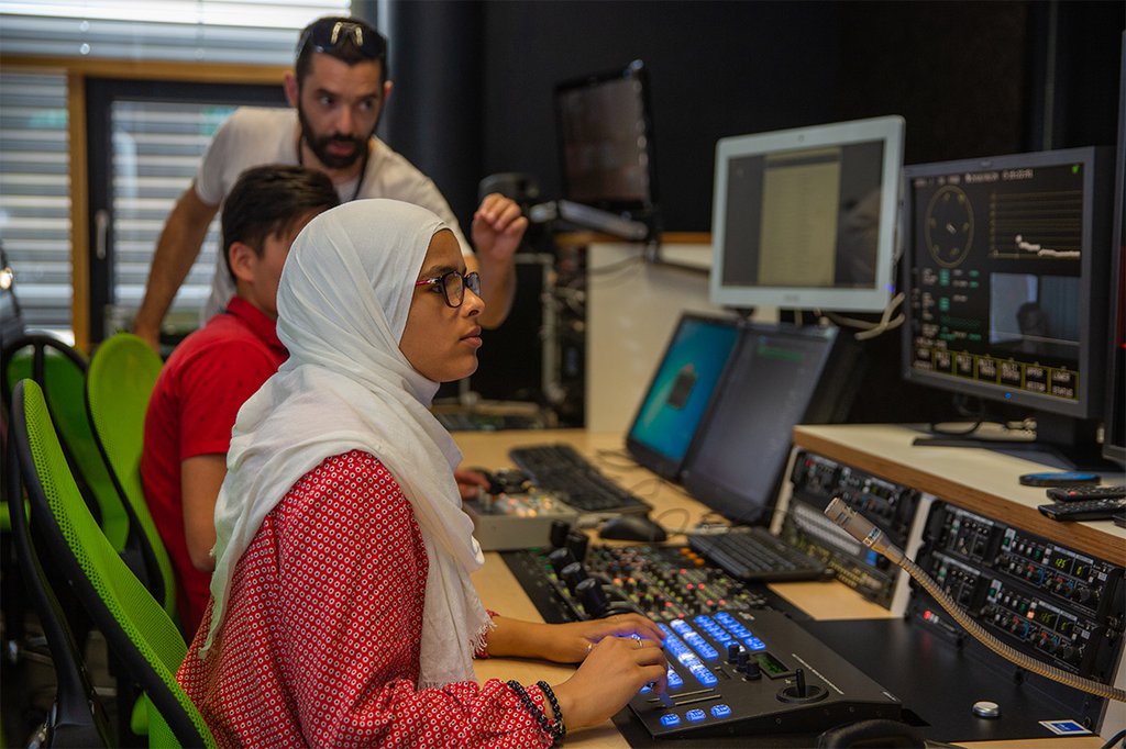 Zwei Jugendliche – eine trägt ein Kopftuch – sitzen im Studio mit verschiedenen Eingabegeräten und Bildschirmen - von der Seite schaut ein Dozent auf den Bildschirm