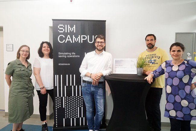 Auf dem Gruppenbild sieht man von links beginnend Lisa Brunhuber und Petra Ganaus stehend bei einem schwarzem SIM CAMPUS Banner. Gleich rechts daneben befindet sich Benjamin Roszipal, Manuel Kaider und Gabriella Szelesi neben einem Stehtisch, ebenfalls stehend.