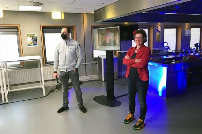 2 speakers, a man and a woman, stand sideways in front of a screen. The man wears an FFP2 mask and holds a microphone.